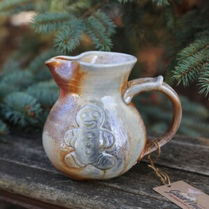 May include: A ceramic pitcher with a gingerbread man design in a light brown glaze with a white and brown glaze. The pitcher has a handle and is sitting on a wooden surface.