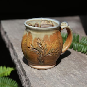 May include: A round ceramic mug with a warm orange and brown speckled glaze. The upper rim features a cream-colored drip pattern. A carved leaf design decorates the front of the mug, which has a sturdy handle on the right side.