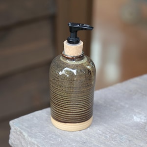 May include: A ceramic soap dispenser with a dark brown, ribbed body and a smooth, light beige base. The top features a natural cork stopper fitted with a black plastic pump mechanism for dispensing liquid soap or lotion in a functional design.