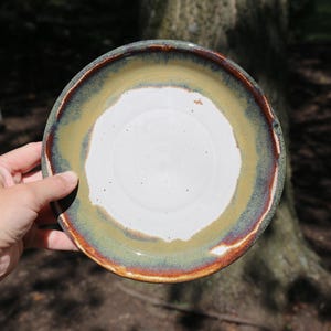 May include: A round ceramic plate featuring a smooth white center surrounded by a wide, textured rim. The rim displays a gradient of earthy olive green and muted brown tones, with a dark, reddish-brown edge that defines the circular shape.
