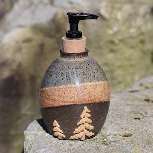 May include: A ceramic soap dispenser with a black pump and a cork stopper. The bottle is a speckled blue-gray at the top, with a tan band and a dark brown base. Two tan pine trees are embossed on the base.