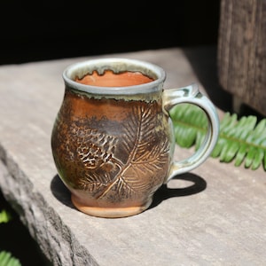May include: A round ceramic mug with a glossy finish, featuring a textured relief of a pinecone and pine needles on the exterior. The mug has a warm brown and orange glaze with a light grey rim and a matching handle, showcasing a handcrafted design.