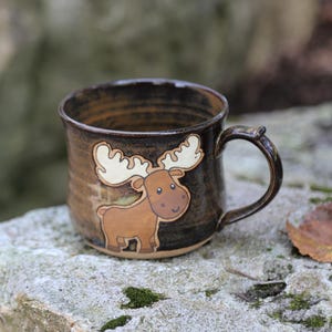 May include: A ceramic mug with a dark brown, glossy glaze featuring a hand-painted illustration of a brown moose with large cream-colored antlers on the front. The mug has a rounded body, a sturdy handle, and a smooth, reflective surface finish.