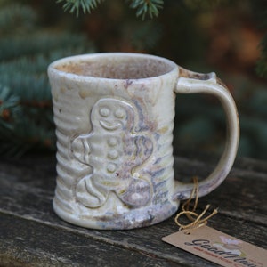 May include: White ceramic mug with a gingerbread man design. The mug has a brown handle and a light brown glaze. The mug is sitting on a wooden surface. "Good Morning" is written on a tag attached to the mug.