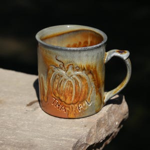 May include: A ceramic mug with a glazed finish in shades of brown, gray, and orange. The mug features a raised, embossed pumpkin design on the front with the word THANKFUL stamped in capital letters below the pumpkin. It has a curved handle.