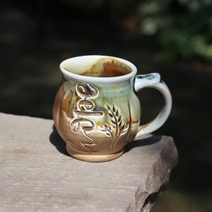 May include: A ceramic mug with a rounded body and a curved handle, featuring a glaze in shades of tan, brown, and sage green. The exterior surface displays an embossed script word that reads "Hope" alongside a carved wheat stalk design.