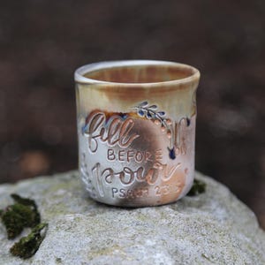 May include: A ceramic tumbler with a light brown and cream glaze. The tumbler features the words "Fill Before You Pour" and "Psalm 23:5" in a carved, cursive font. Decorative floral accents are also present.