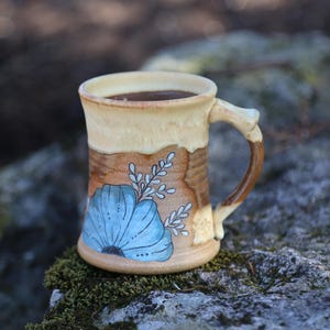May include: Handmade ceramic mug with a light brown exterior and a dark brown handle. The mug features a blue floral design with white accents. The interior is filled with a dark liquid, likely coffee or tea. The mug is sitting on a mossy rock.