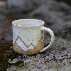 May include: A ceramic mug with a speckled off-white glaze on the upper portion and a textured, light brown clay base. The lower section features a carved mountain range design with white accents, a rounded handle, and a wide, open circular rim.