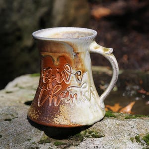 May include: A ceramic mug with a flared base and a handle, featuring a two-tone glaze of cream and earthy brown. The words "Give thanks" are embossed in a cursive script on the front, with the brown glaze highlighting the textured lettering.