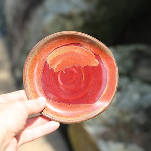 May include: A small, round ceramic plate with a vibrant, glossy finish. The plate features a gradient of colors, transitioning from a deep red center to a warm orange rim. The plate is held in a hand, showcasing its size and design.