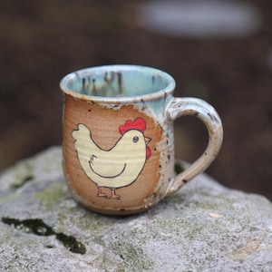 May include: A handmade ceramic mug with a whimsical chicken design. The mug features a light brown exterior with a light blue interior and handle. A yellow chicken with a red comb is painted on the side. Perfect for coffee or tea.