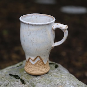 May include: A handmade ceramic mug with a cream-colored glaze and speckled finish. The mug features a mountain design at the base and a curved handle. The mug is approximately 4 inches tall.