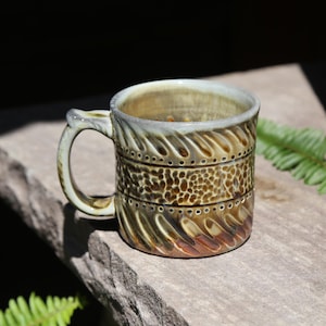 May include: A ceramic mug with a handle, featuring a multi-textured exterior with a central band of honeycomb-like indentations flanked by rows of small dots and diagonal carved grooves, finished in a glossy brown and cream glaze.