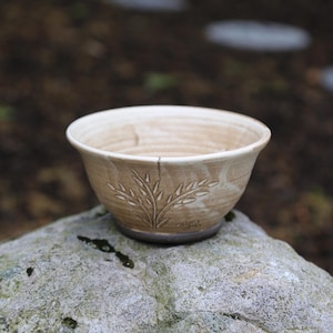 May include: A handmade ceramic bowl with a light brown exterior and a cream-colored interior. The bowl features an embossed leaf design on the side. The bowl is sitting on a gray rock. This bowl is perfect for serving food.