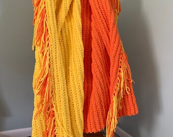 May include: Two crocheted blankets with fringe, one yellow and one orange, draped over a wooden stool.