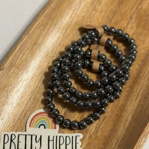 Könnte beinhalten: Vier schwarze Perlenarmbänder, die auf einer Holzoberfläche gestapelt sind. Die Armbänder bestehen aus kleinen, runden Perlen. Der Text "Pretty Hippie" ist auf einem Aufkleber im Vordergrund sichtbar.