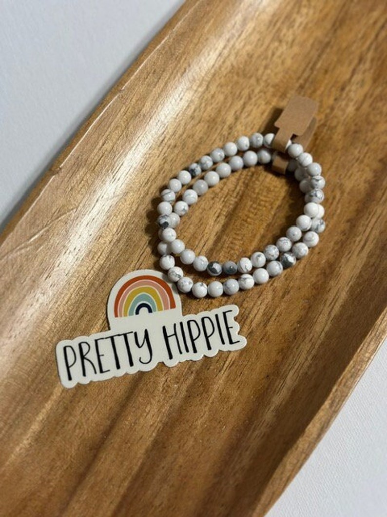 Puede incluir: Un brazalete de cuentas blancas y grises con una pegatina que dice "Pretty Hippie" y un dise&ntilde;o de arco&iacute;ris.