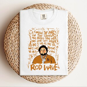 K&ouml;nnte beinhalten: Wei&szlig;es T-Shirt mit einer orangefarbenen Grafik von Rod Wave und Text. Der Text enth&auml;lt den Satz &bdquo;Ich habe die Party des Jahrhunderts geschmissen&ldquo; und &bdquo;Rod Wave&ldquo;. Das T-Shirt ist auf einer gewebten, kreisf&ouml;rmigen Oberfl&auml;che gefaltet.