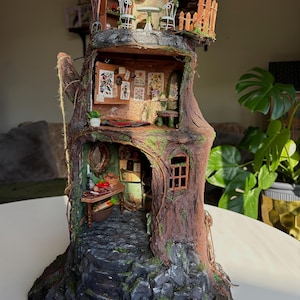 May include: A detailed miniature treehouse, constructed from wood and decorated with moss and vines. It features multiple levels, a small balcony with chairs, and a cosy interior with miniature furniture. A stone pathway leads to the entrance. A whimsical decorative item.