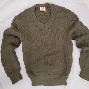 Könnte beinhalten: Olivgrüner V-Ausschnitt-Pullover mit Rippstrickstruktur. Der langärmlige Pullover hat ein klassisches Design mit geripptem Saum und Bündchen. Ein kleines Etikett ist in der Nähe des Halsausschnitts sichtbar. Dieser Pullover ist für den Freizeitgebrauch geeignet.