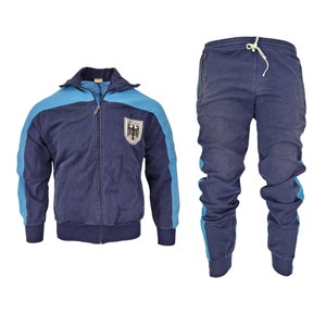 May include: A navy blue tracksuit with light blue accents. The jacket has a zip fastening and a German eagle logo on the chest. The trousers have a drawstring waistband and side pockets.