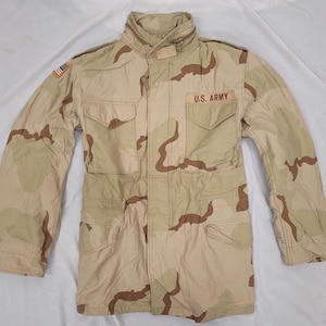 May include: A desert camouflage military jacket in tan and brown, featuring the text "U.S. ARMY" on the chest. It has multiple pockets, a high collar, and a small American flag patch. The jacket is made of a robust material.