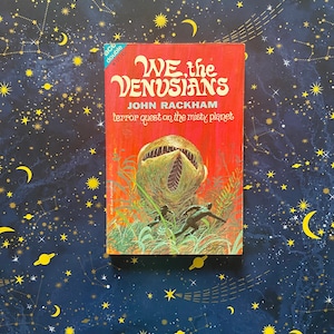 Könnte beinhalten: Ein rotes Taschenbuch mit dem Titel "WE, the VENUSIANS" von John Rackham. Der Umschlag zeigt eine Illustration einer großen, zahnigen Pflanze, von der zwei Figuren weglaufen.