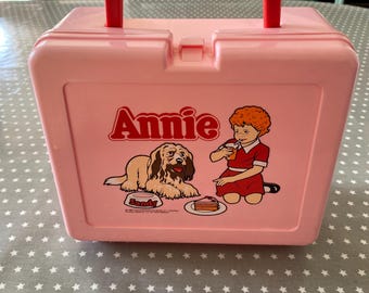 May include: A pink plastic lunchbox with a red handle and clasp. The lunchbox has the name "Annie" in red, with illustrations of a girl, a dog, and cake. It rests on a grey tablecloth with white stars.