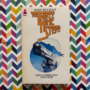 May include: A vintage paperback book titled "The Thirty Nine Steps" by John Buchan. The cover shows a train, a plane, and a hand grasping a train. The book is set against a colorful chevron pattern background.