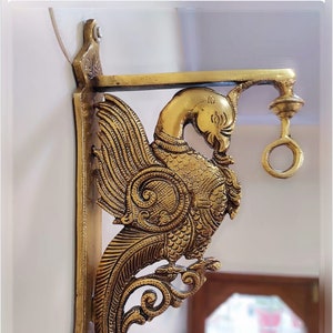 Può includere: Un supporto da parete in metallo dorato a forma di uccello stilizzato con dettagli intricati. Il supporto ha un anello attaccato alla parte superiore per appendere oggetti.