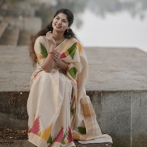 May include: A woman wearing a white and cream saree with a colorful geometric pattern. The saree has a green, yellow, and red border. She is sitting on a concrete step with a body of water in the background.