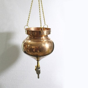 May include: A copper hanging pot with a brass spout and a gold chain. The pot is hanging from a wooden beam.