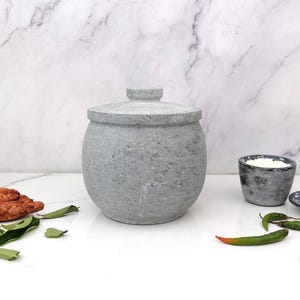 May include: A gray stone jar with a lid, a small bowl, and a small plate. The jar is round and textured. Green chili peppers and leaves are scattered around the objects. The background is a white marble.