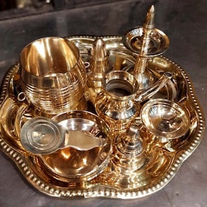 Op de afbeelding: Een set van zeven gouden metalen ceremoni&euml;le voorwerpen, waaronder een kleine kan, een kom, een beker, een kleine pot, een deksel en een decoratief stuk, allemaal gerangschikt op een gouden metalen dienblad met een parelrand.