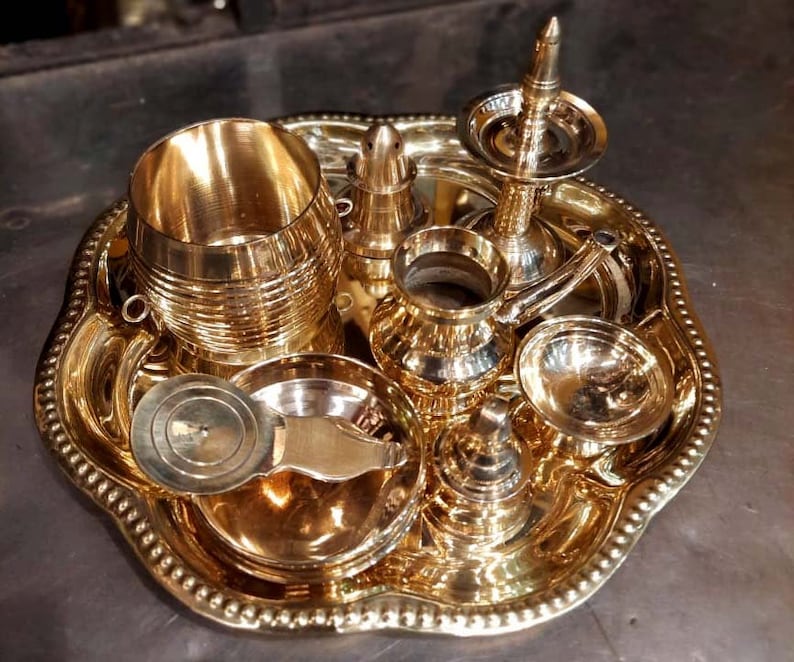Op de afbeelding: Een set van zeven gouden metalen ceremoni&euml;le voorwerpen, waaronder een kleine kan, een kom, een beker, een kleine pot, een deksel en een decoratief stuk, allemaal gerangschikt op een gouden metalen dienblad met een parelrand.