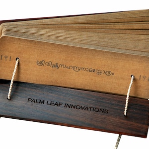 Puede incluir: Un libro manuscrito de hojas de palma marrón con una cubierta de madera oscura. El libro está atado con una cuerda y tiene el texto "PALM LEAF INNOVATIONS" en la cubierta.