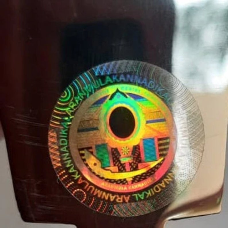 May include: A holographic sticker with a circular design featuring a colorful mandala with a black center and the text "ARAMULA KANNADIKA" repeated around the edge.