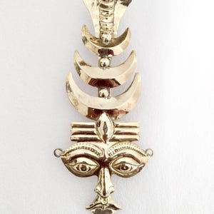 May include: A gold metal wall hanging featuring a stylized face with a cobra hood and crescent moons above. The face has large eyes and a detailed mouth.