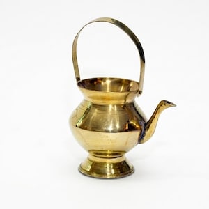May include: A polished brass vessel featuring a rounded body, a wide circular opening, and a curved spout. The container includes a thin, arched metal handle attached to the sides, resting on a flared pedestal base with subtle horizontal line details.