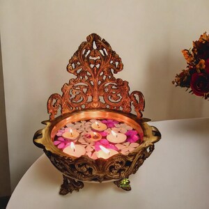 May include: Ornate brass bowl with a decorative back, filled with water, floating candles, and flower petals. The bowl has intricate carvings and stands on three legs. The candles are lit, creating a warm, inviting glow.