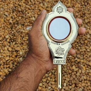 Op de afbeelding: Een gouden handspiegel met een houten handvat. De spiegel heeft een ronde, reflecterende oppervlak en een decoratief ontwerp rond de rand. Het handvat is gegraveerd met de tekst "Aranmula Kaniyadi".