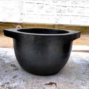 May include: A black, rounded bowl with a flat, rectangular handle on each side. The bowl has a smooth, matte finish and appears to be made of stone or a similar material. The bowl is sitting on a concrete surface.