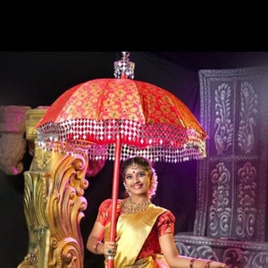 May include: A woman in a gold and red saree holds a red and gold umbrella with a silver trim. The umbrella is decorated with small, shiny, silver ornaments. The woman is smiling and looking at the camera.