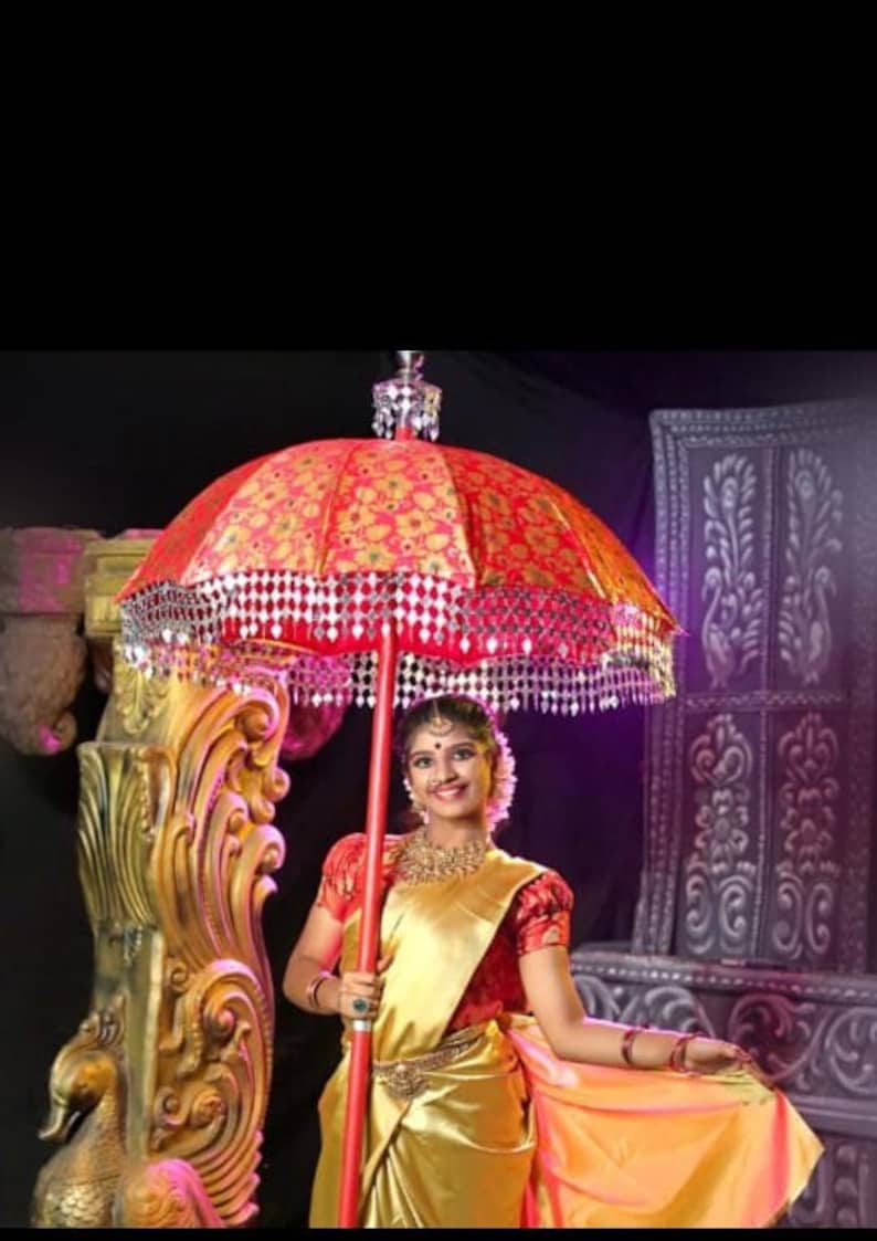 May include: A woman in a gold and red saree holds a red and gold umbrella with a silver trim. The umbrella is decorated with small, shiny, silver ornaments. The woman is smiling and looking at the camera.