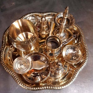 Op de afbeelding: Een set van acht gouden metalen ceremoni&euml;le voorwerpen op een decoratief dienblad. De voorwerpen omvatten een ​​kleine kan, een kom, een kopje, een kleine pot, een container met deksel en drie kleine kommen.