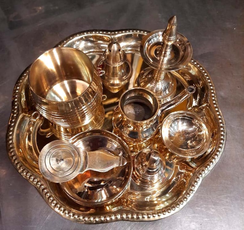 Op de afbeelding: Een set van acht gouden metalen ceremoni&euml;le voorwerpen op een decoratief dienblad. De voorwerpen omvatten een ​​kleine kan, een kom, een kopje, een kleine pot, een container met deksel en drie kleine kommen.