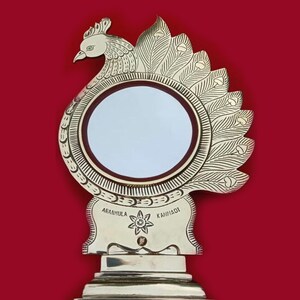 May include: A gold-colored peacock-shaped decorative mirror with a round, white reflective surface. The mirror has a base with the words "AARANMULA KAMNADI" inscribed on it.