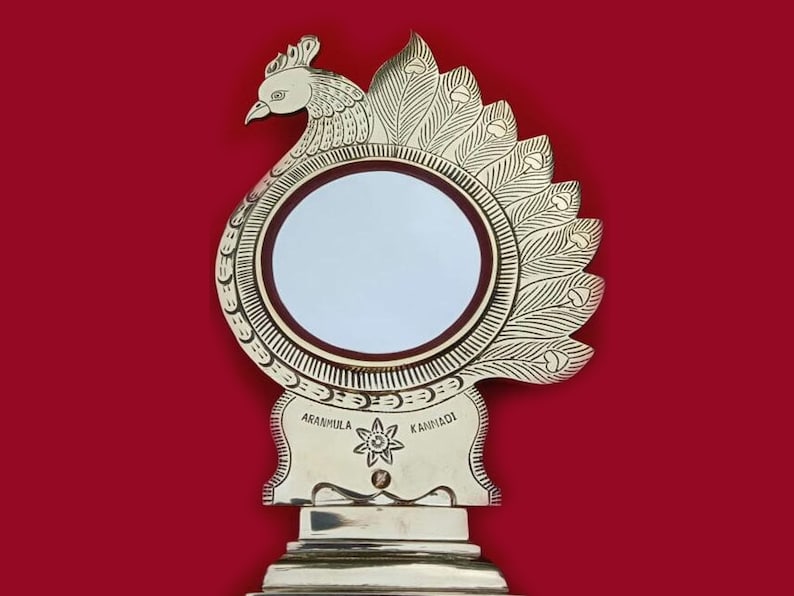 May include: A gold-colored peacock-shaped decorative mirror with a round, white reflective surface. The mirror has a base with the words "AARANMULA KAMNADI" inscribed on it.