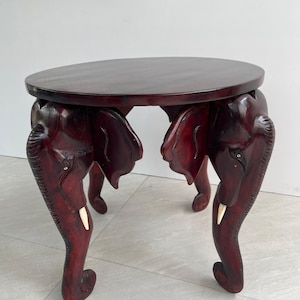 Puede incluir: Una mesa de madera de color rojo oscuro con tapa redonda y cuatro patas talladas en forma de elefantes. Los elefantes tienen colmillos de marfil y tallas detalladas.